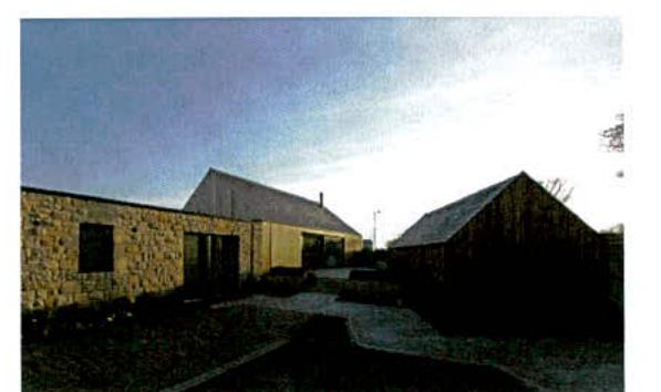 A photograph showing a rural property with existing agricultural buildings and a new single-story stone structure with a driveway leading to it.