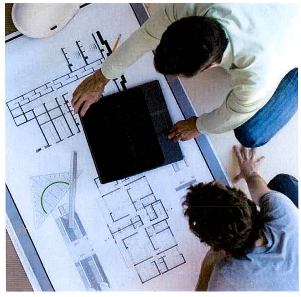 Two individuals are reviewing architectural floor plans and elevations spread out on a table with drafting tools.
