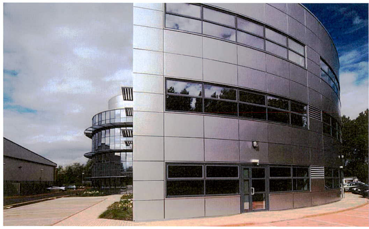 A photograph showing the exterior of a modern commercial building with a curved grey facade, large windows, and an external spiral staircase structure.