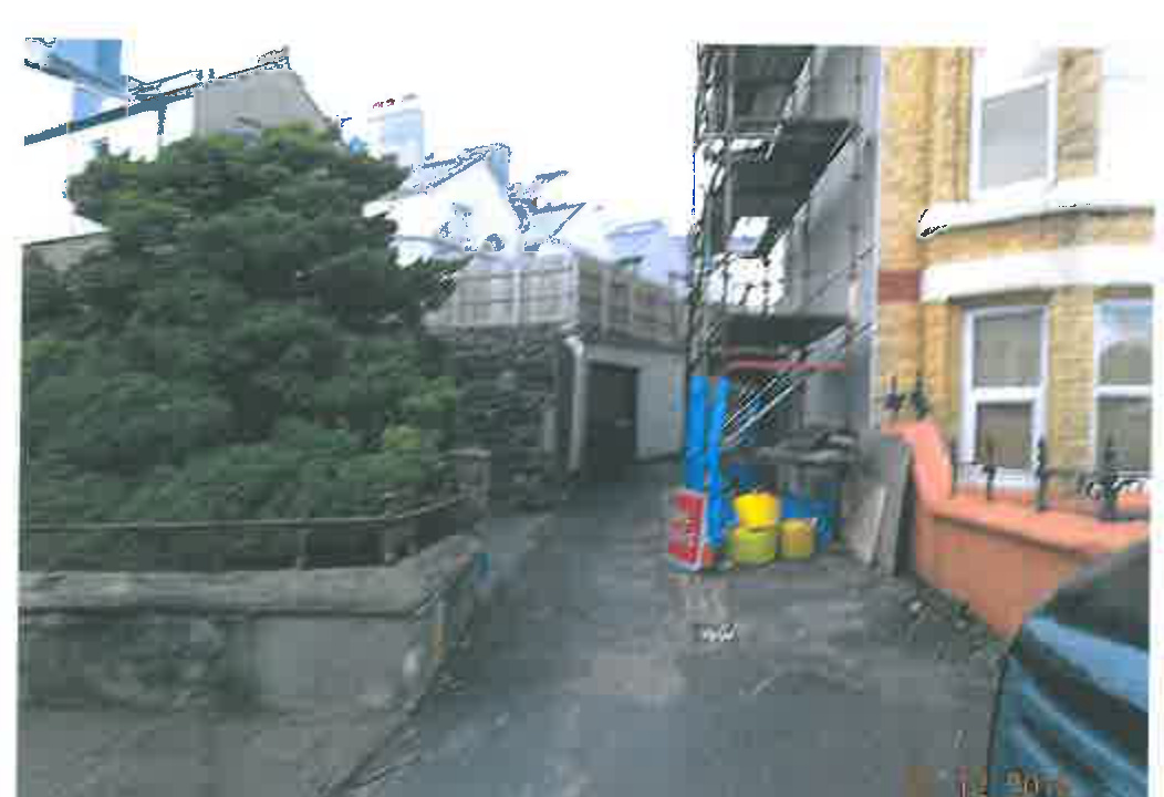 A photograph showing the rear or side exterior of a residential property, featuring a brick house, a garage or outbuilding, and a paved driveway area with some construction materials.