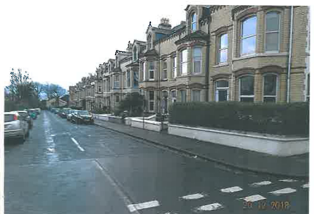 A street-level photograph showing a row of terraced houses along a wet road with parked cars and a low boundary wall.