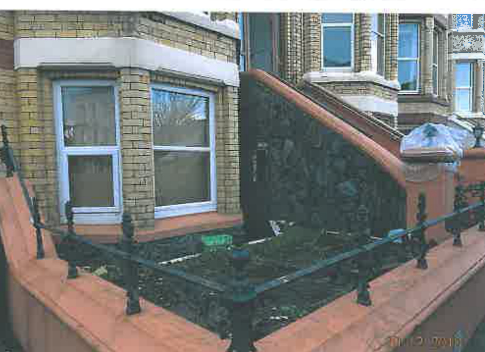 A photograph showing the front exterior of a brick residential property featuring a bay window, steps leading to the entrance, and a boundary wall with railings.