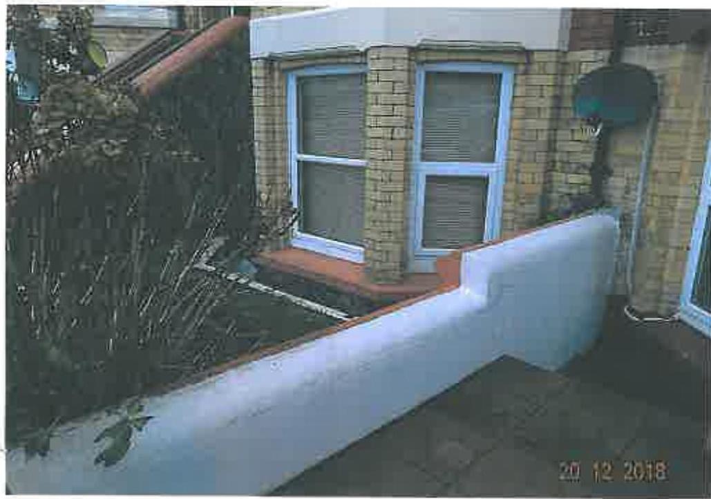 A photograph showing the exterior brick wall of a single-story building with white-framed windows and a low white boundary wall in the foreground.