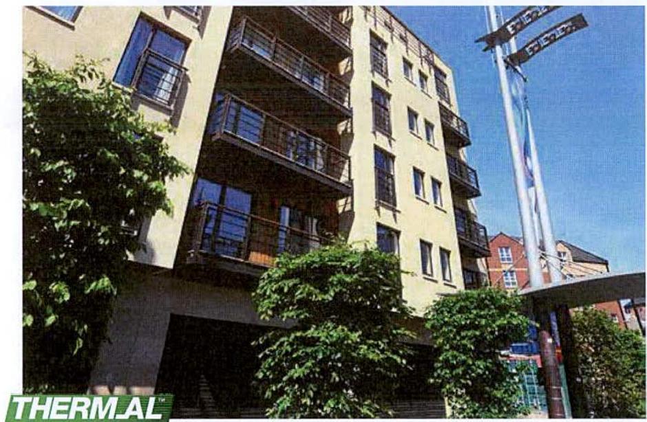A low-angle photograph of a multi-story building with balconies and large windows, partially obscured by trees in the foreground.