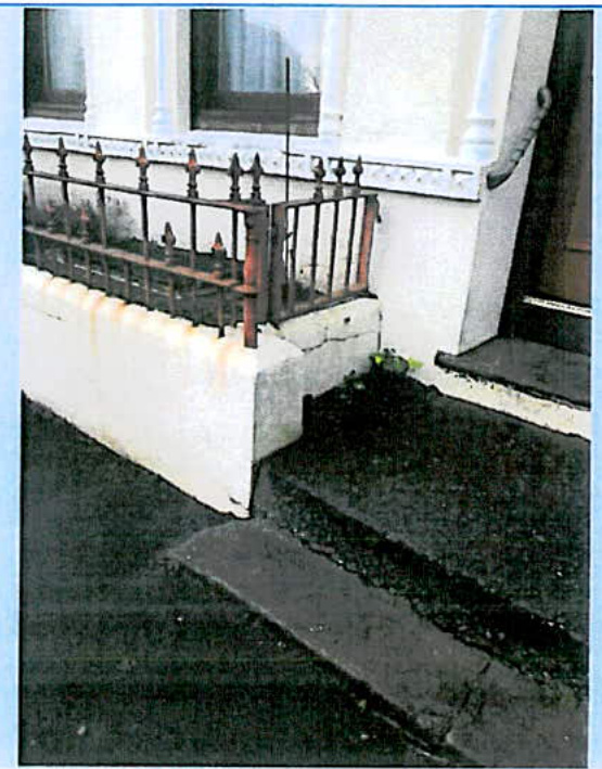 A photograph showing the exterior entrance of a building featuring stone steps leading to a door and a raised area protected by a black metal railing.