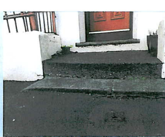 A close-up photograph showing concrete steps leading up to a red front door, flanked by white walls and a black railing.