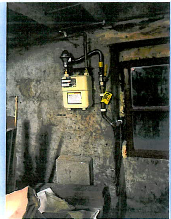 Interior photograph showing a yellow gas meter mounted on a rough wall with black piping and a window frame to the right.