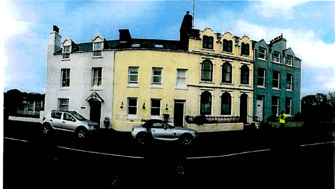 A photograph showing a row of terraced buildings painted in white, yellow, and blue, with cars parked on the street in front.