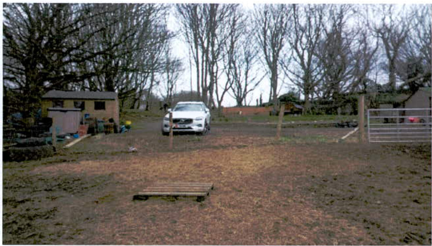 This photograph depicts a rural paddock or yard with muddy ground, featuring a white car parked centrally, a small wooden shed to the left, and a metal gate to the right.