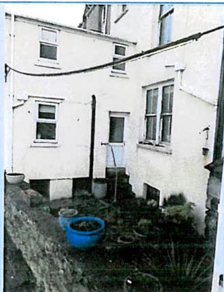 A grainy photograph showing the rear exterior of a white-washed residential building with a small patio area containing a blue tub and potted plants.