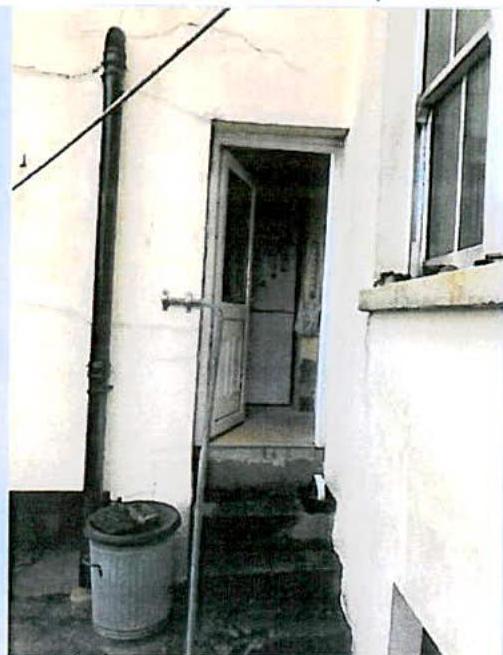 A photograph showing the side exterior of a white building with an open door, steps leading up to it, a window, and a vertical drainpipe.