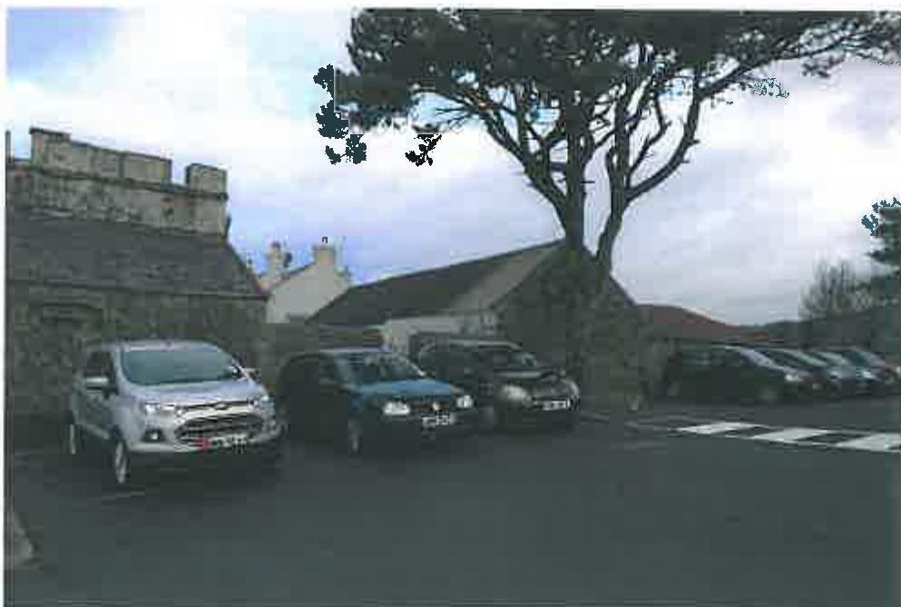 A photograph showing a paved parking area with several cars parked in front of traditional stone buildings and a large tree.