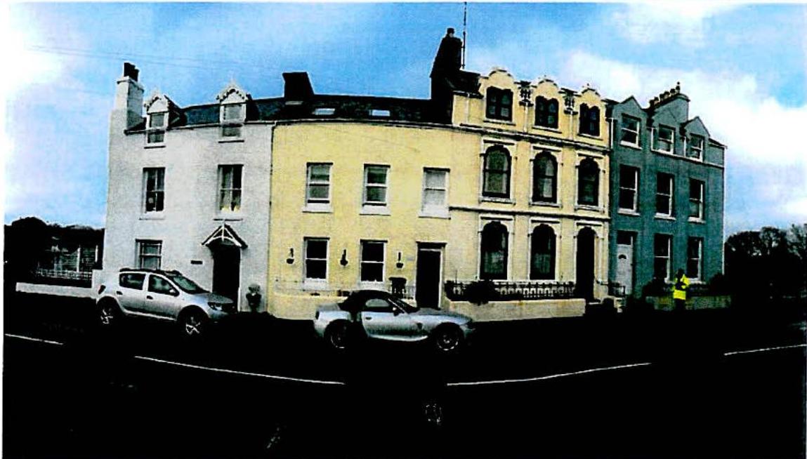 A street-level photograph showing a row of terraced buildings, including a central cream-colored house and adjacent white and blue structures, with cars parked on the street.