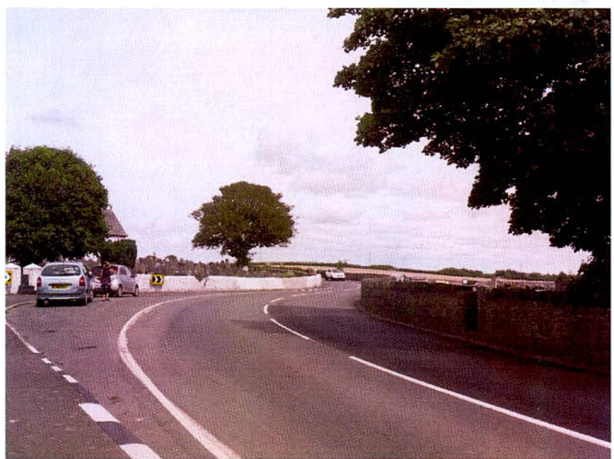 A photograph showing a curved rural road with parked cars on the left and stone walls and trees on the right.