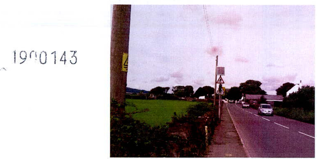 A photograph showing a rural road scene with a large green field on the left and vehicles driving on the road to the right.