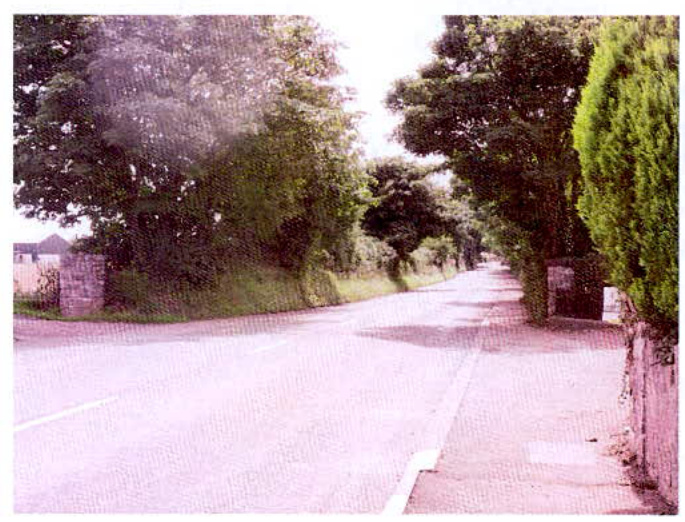 A grainy photograph showing a paved rural road flanked by trees and stone walls, likely illustrating the existing access route to the site.