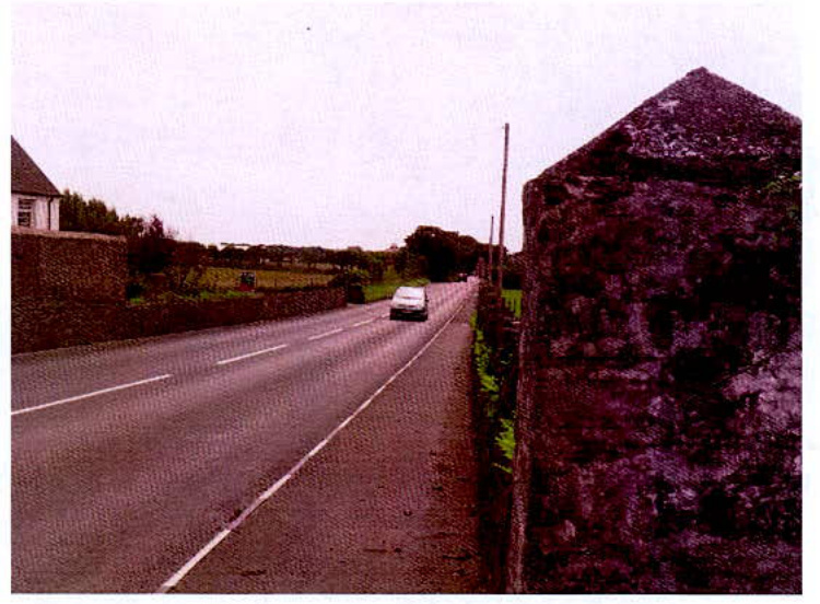A photograph showing a rural road scene with a car driving away, flanked by stone walls and hedgerows, likely depicting the site access or surroundings.