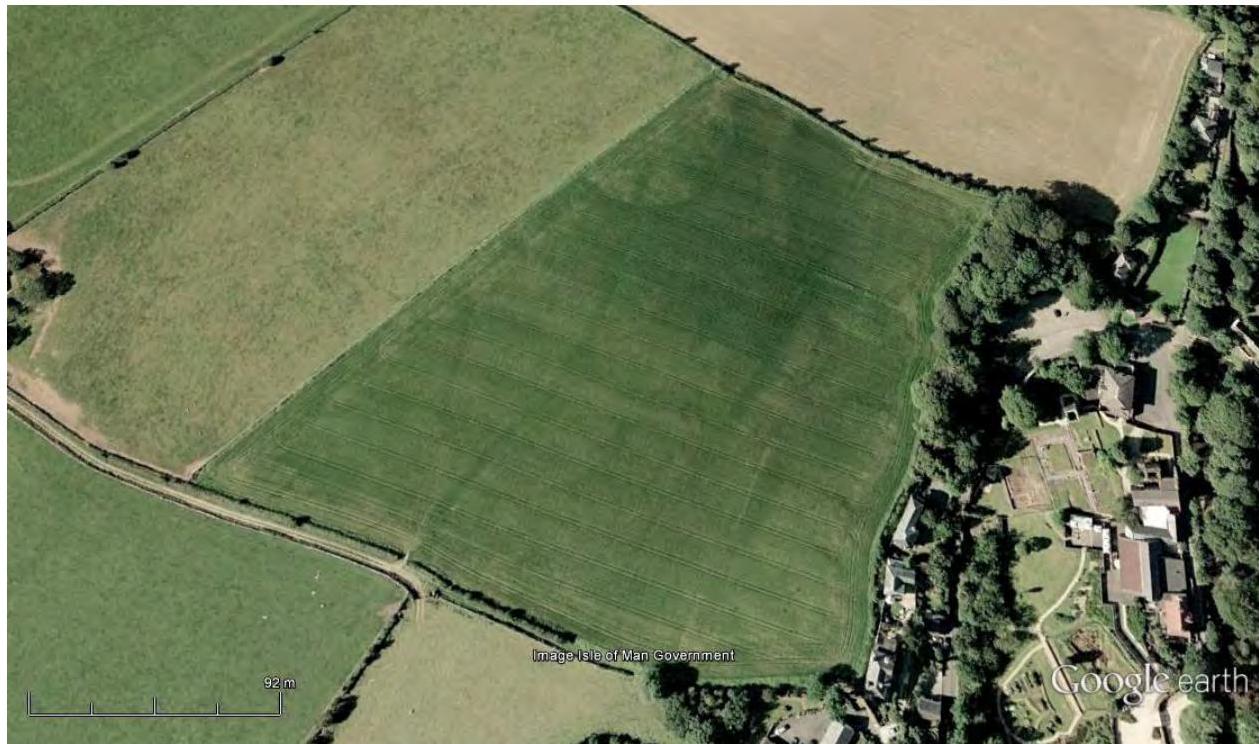 An aerial satellite view of a large green agricultural field bordered by hedgerows, with residential properties visible on the right side.
