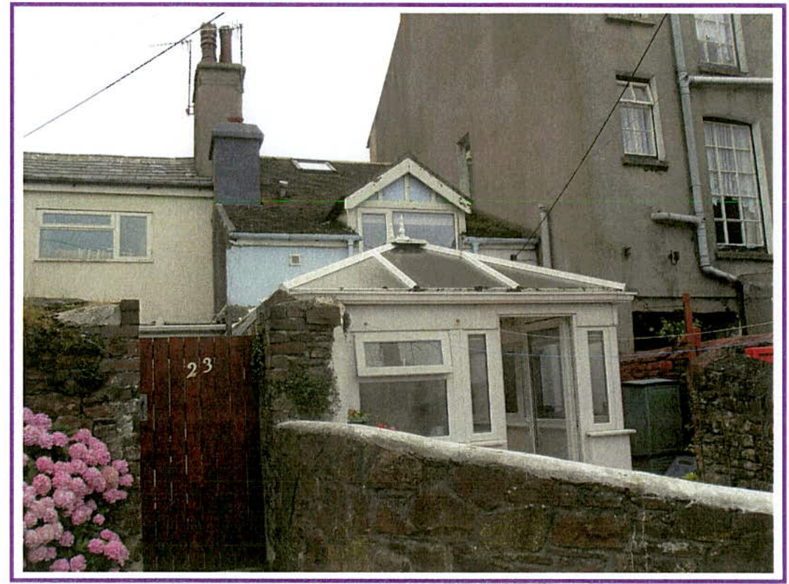 A photograph showing the exterior of a residential property featuring a white conservatory, a wooden gate with the number 23, and adjacent stone buildings.