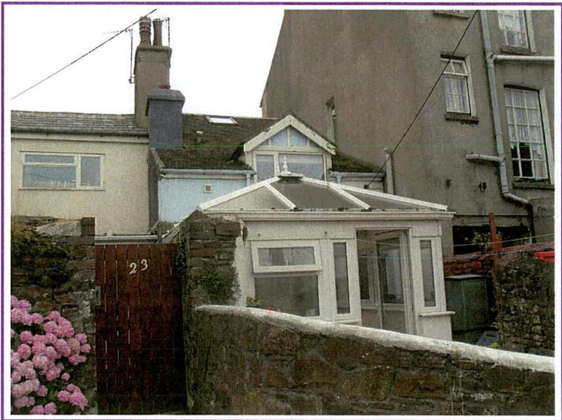 A photograph showing the exterior of a residential property featuring a white conservatory extension, stone boundary walls, and a wooden gate with the number 23.