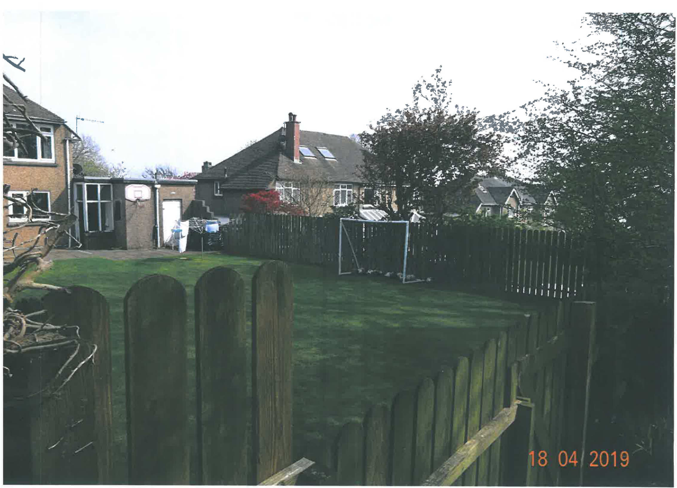 A photograph showing a residential rear garden with a wooden fence, grass lawn, and a soccer goal. Neighboring houses and trees are visible in the background.