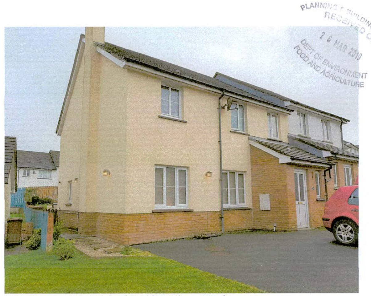 A photograph showing the side elevation of a two-story cream-colored house with a brick base, featuring a driveway and a parked red car.