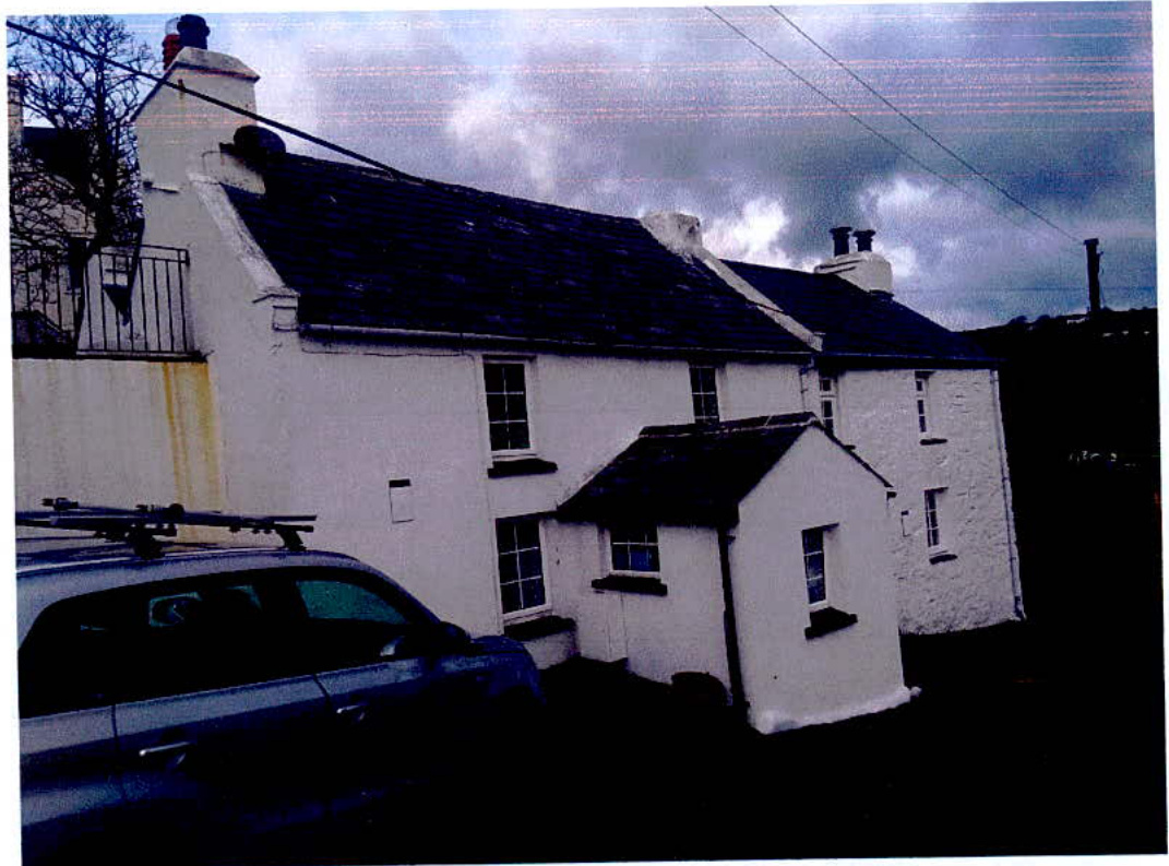A photograph showing the front elevation of a white, two-story terraced house with a small single-story extension attached. A car is parked in the foreground on the left side.