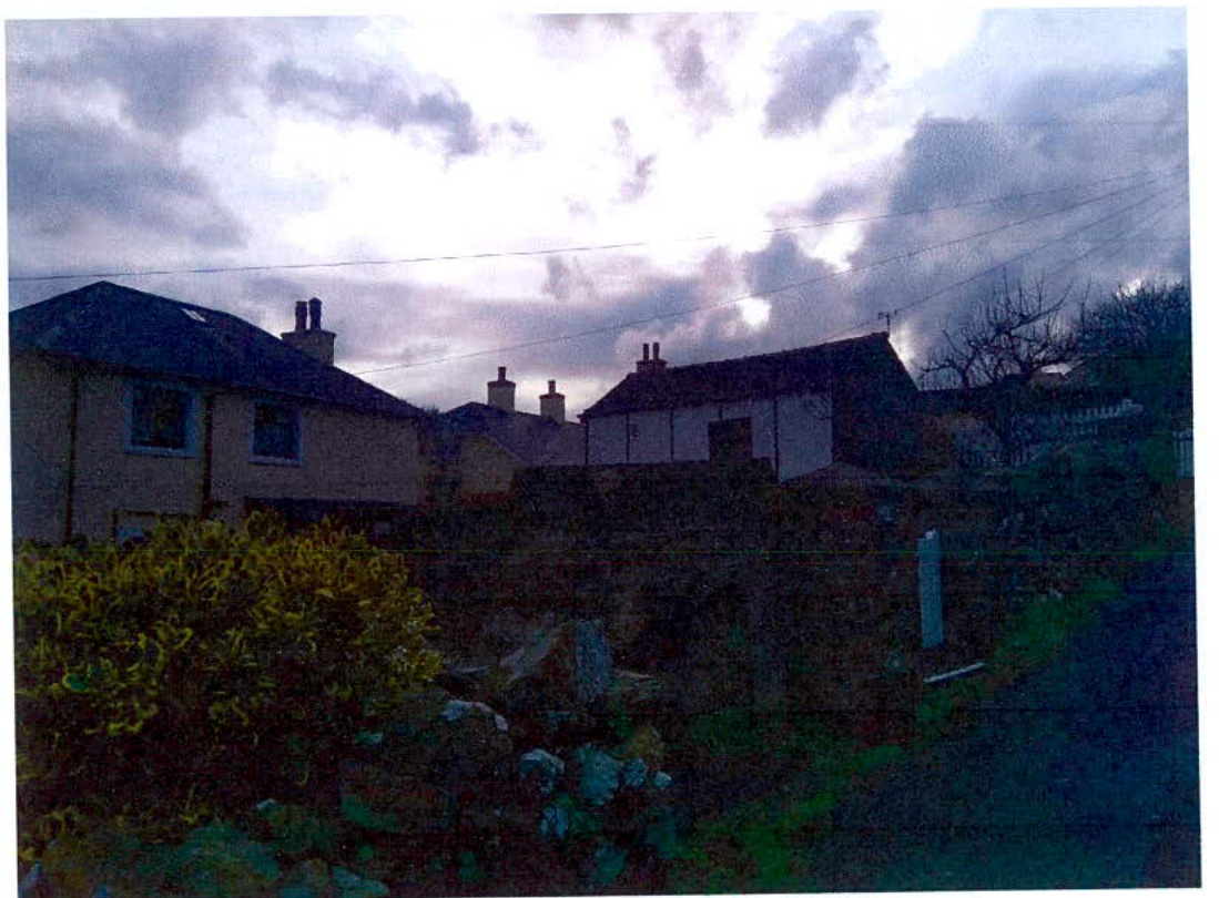 A grainy photograph showing a row of residential buildings with stone walls and vegetation in the foreground, likely depicting the existing site context.