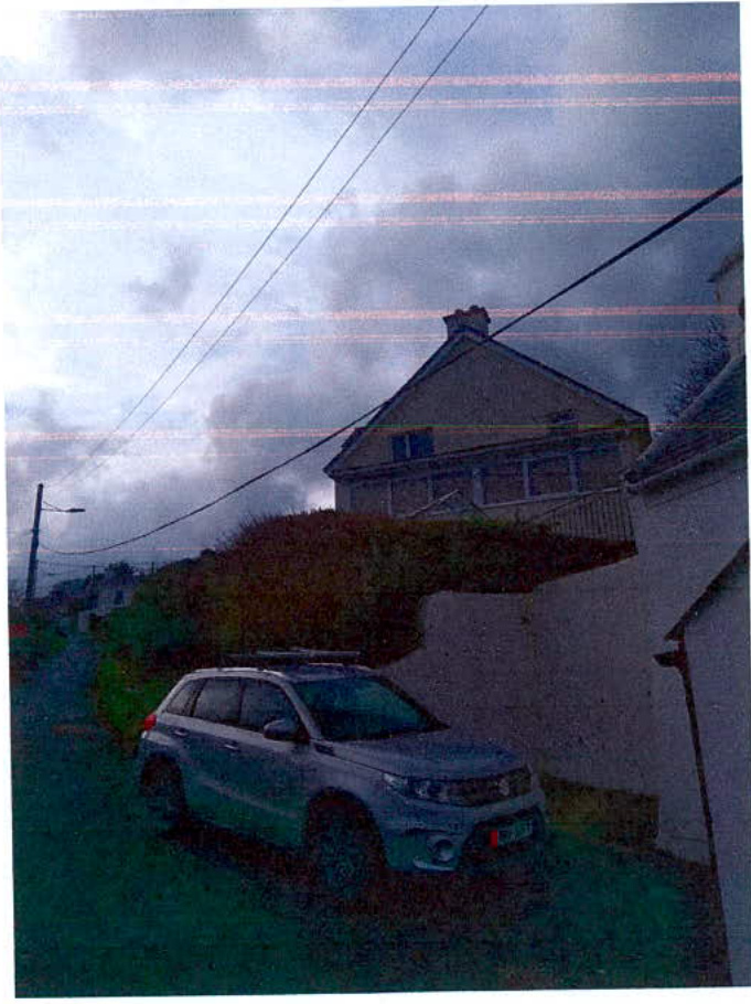 A grainy photograph showing the exterior of a detached house situated on a slope, with a car parked in the foreground.