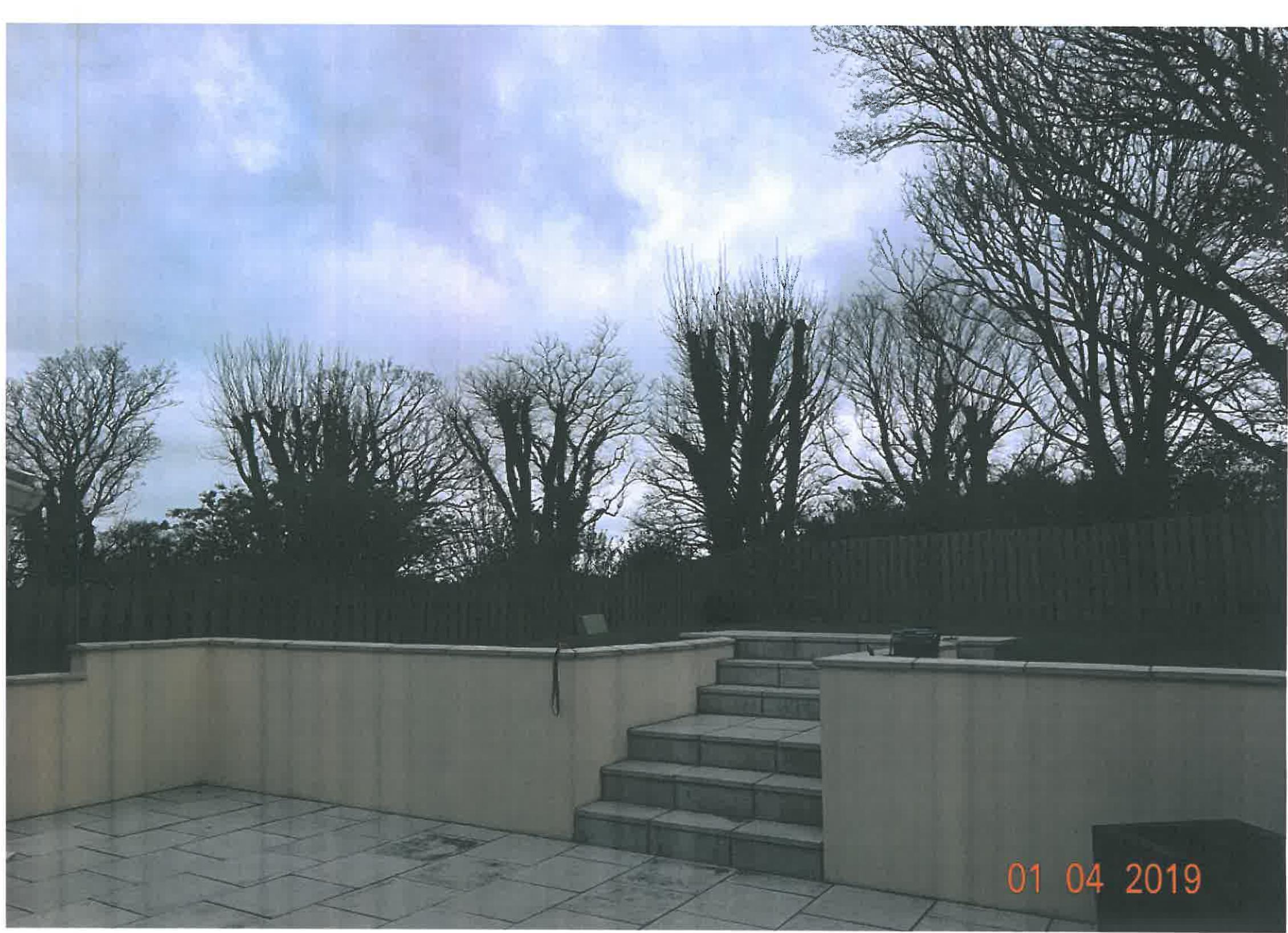A photograph showing a paved patio area with a low retaining wall and steps leading up to a wooden fence and trees.