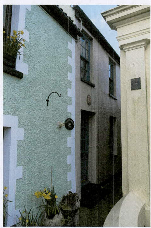 A photograph showing the exterior facade of a terraced building with light green render and white quoins, featuring a window box with yellow flowers.