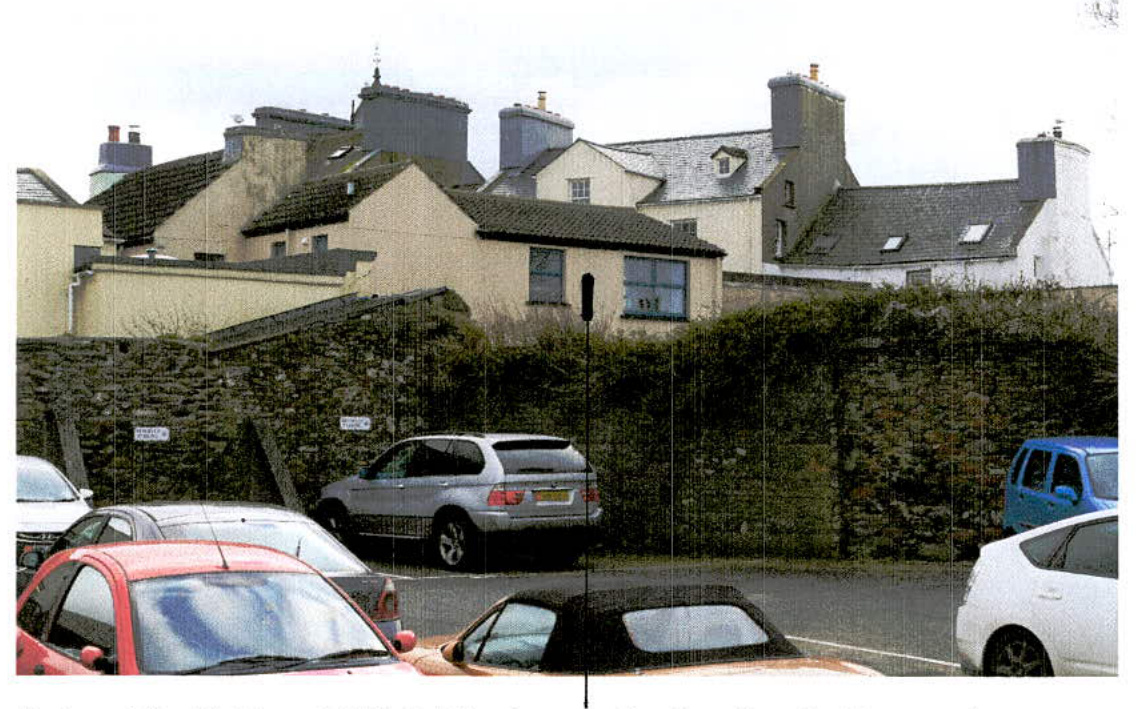 A street-level photograph showing a row of residential buildings behind a stone wall and hedge, with cars parked in the foreground.