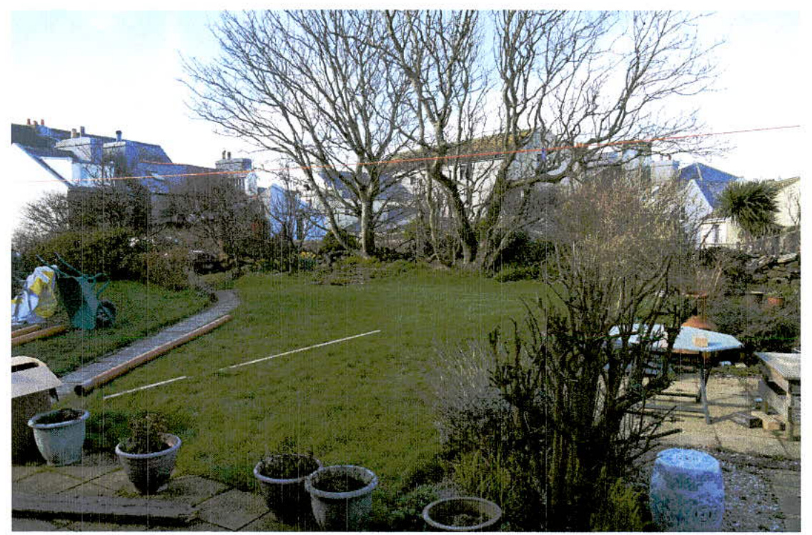 A photograph showing a residential garden with a large lawn, mature trees, and patio furniture, with neighboring houses visible in the background.