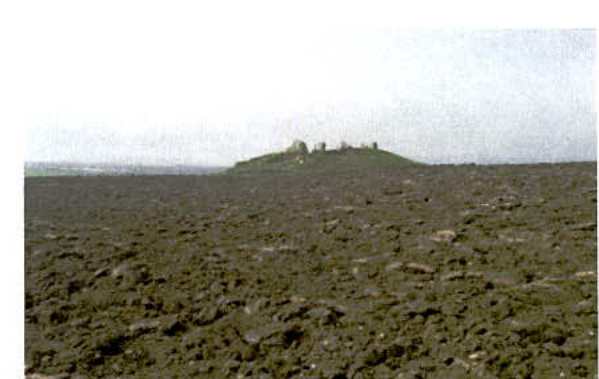 A grainy photograph showing a rough, ploughed field in the foreground with a hill topped by stone ruins visible in the distance.