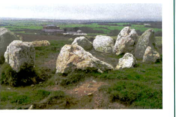 A photograph showing a grassy field with large standing stones in the foreground and a rural landscape with a distant building in the background.