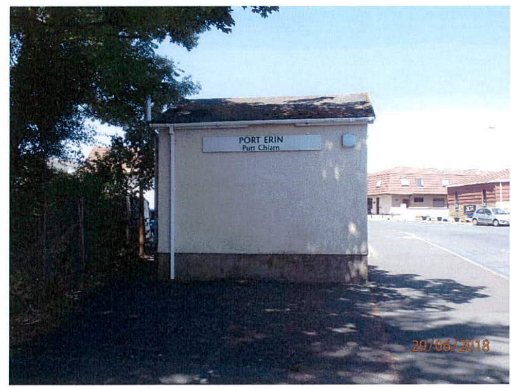 A photograph of a small, single-story building with a sign reading 'PORT ERIN' and 'Purt Chiarn', situated next to a road with trees and other buildings in the background.