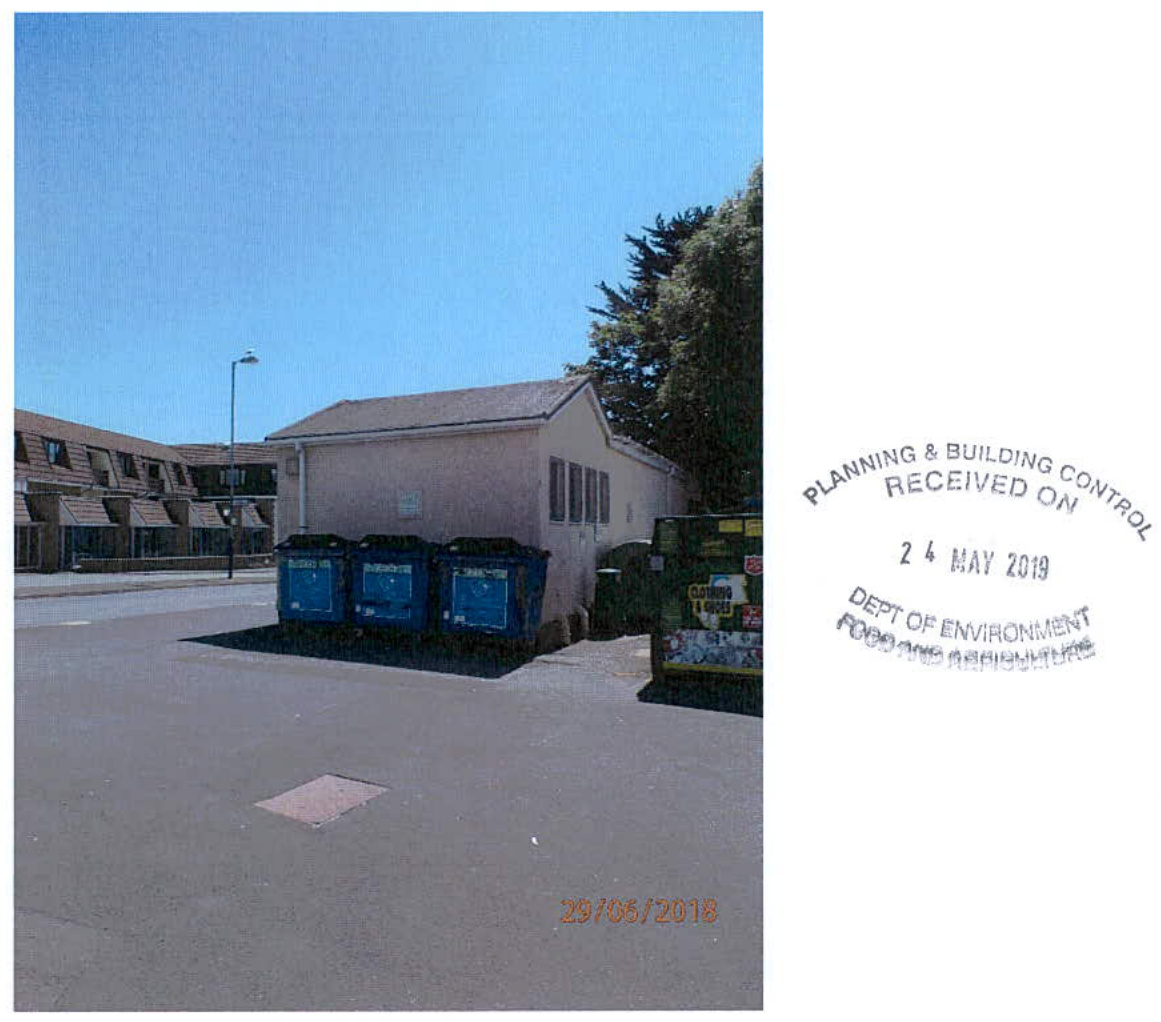 A photograph showing a single-story building with blue recycling bins in the foreground and terraced housing in the background, likely the subject of the heritage centre conversion application.