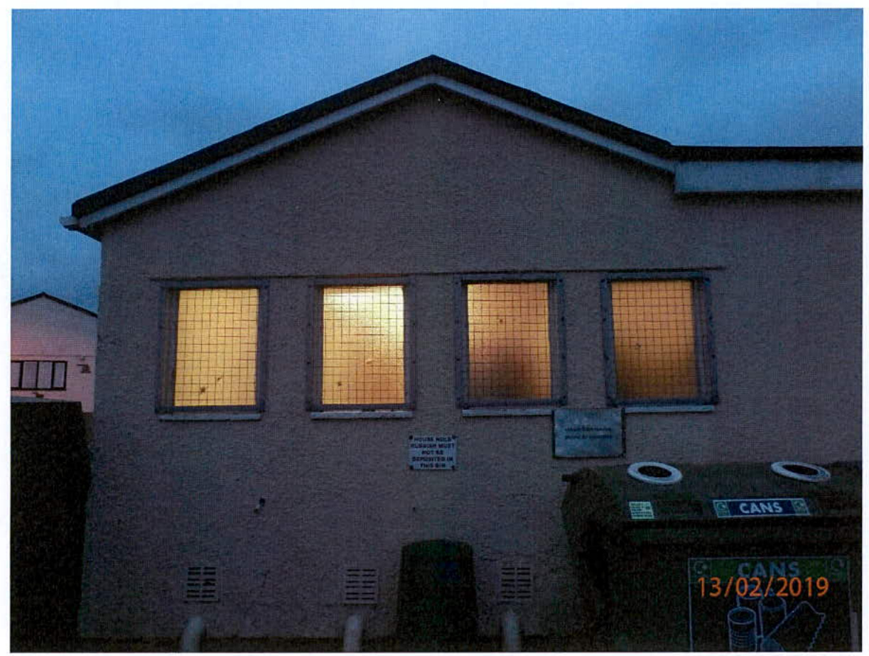 A photograph showing the exterior facade of a single-story building at dusk, featuring four large windows with wire mesh and recycling bins in the foreground.