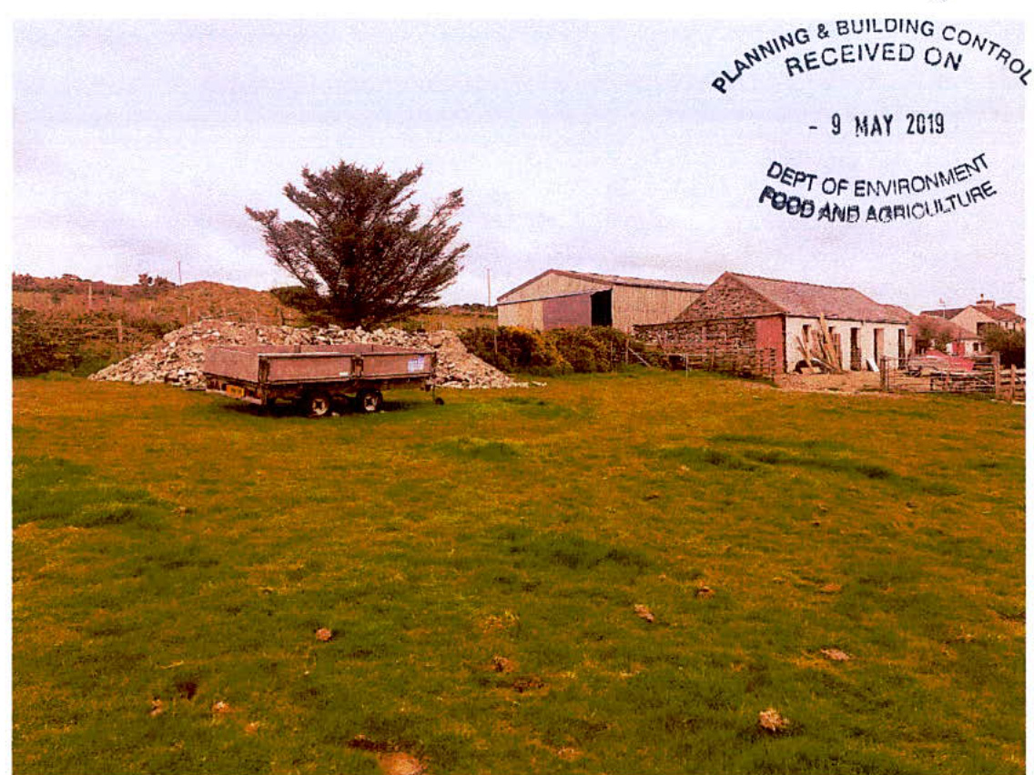 A photograph of a rural site showing a grassy field, a trailer with stones, and existing agricultural buildings in the background.