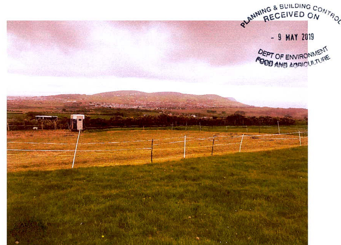 A photograph showing a rural grassy field with electric fencing and a small white shelter, set against a backdrop of rolling hills and a distant settlement.
