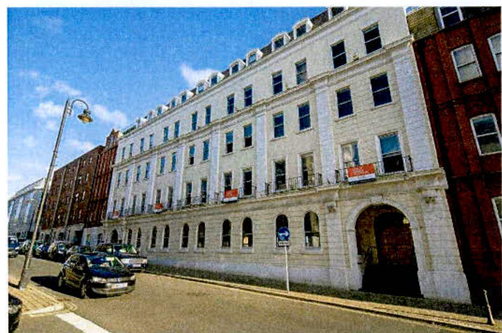 A street-level photograph showing a large, white, multi-story building with arched ground-floor windows and rectangular upper windows, situated on a paved road with parked cars.