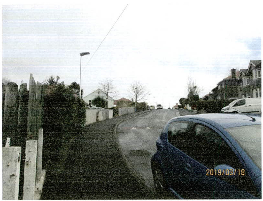 A grainy photograph showing a residential street curving uphill with a blue car parked in the foreground and houses lining the road.