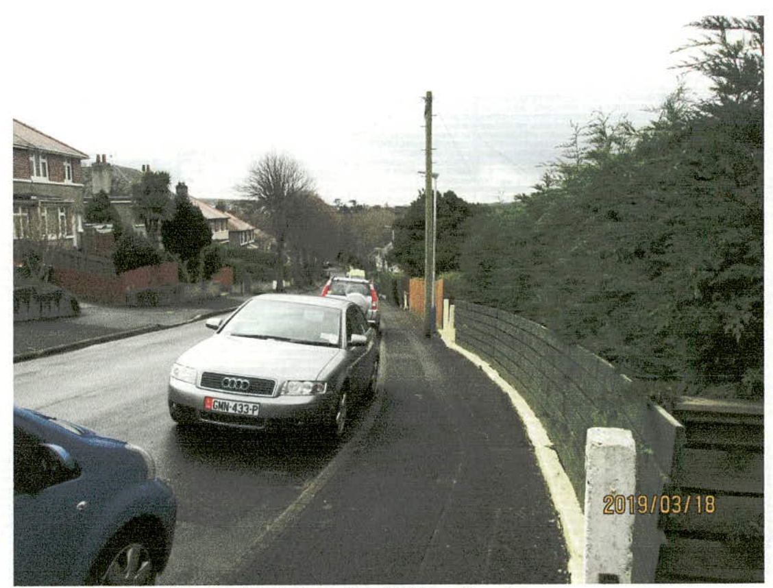 A street-level photograph showing cars parked along a narrow residential road with a sidewalk and fence on the right.