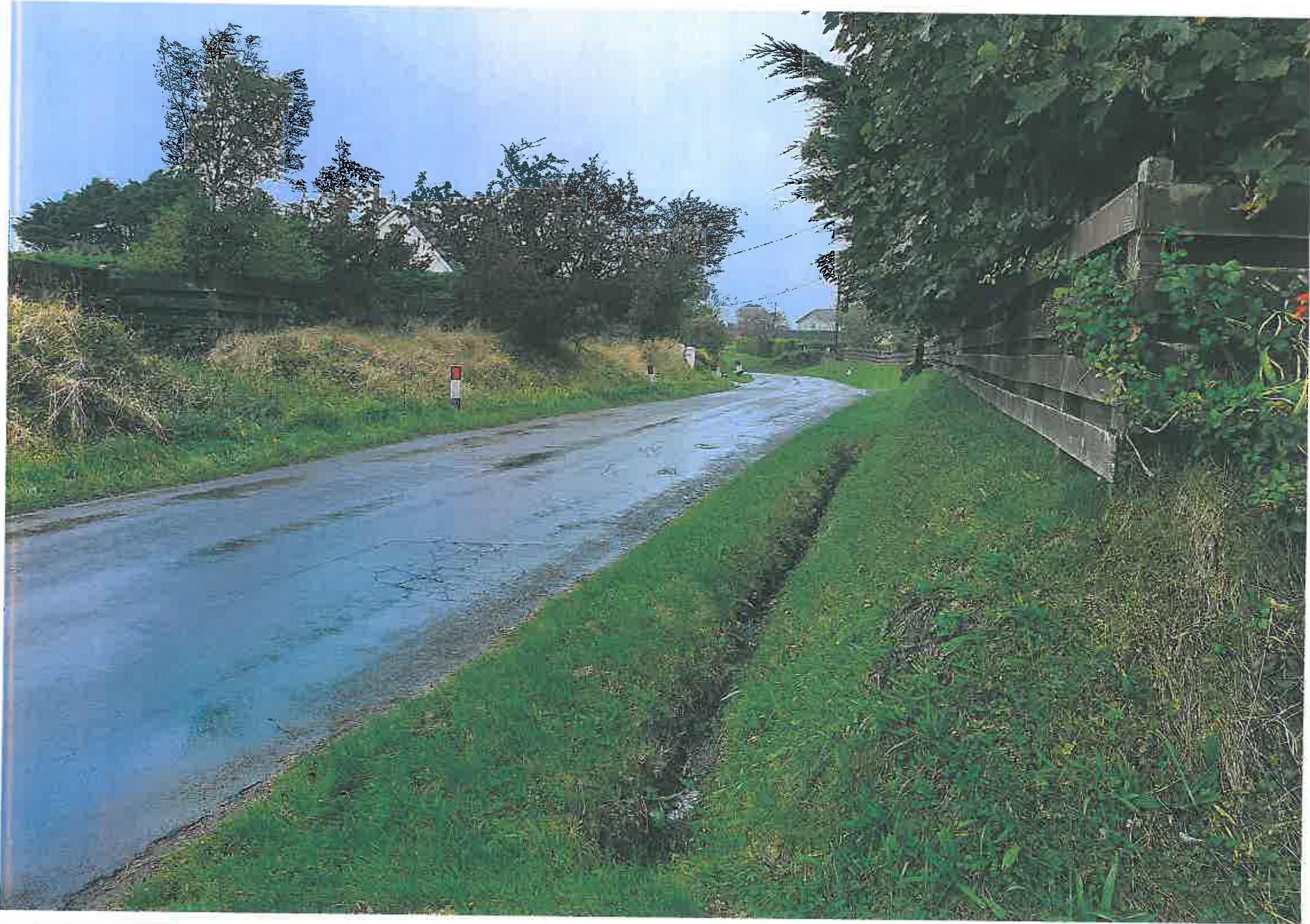 A photograph showing a wet, paved rural lane bordered by grassy banks and wooden fencing.