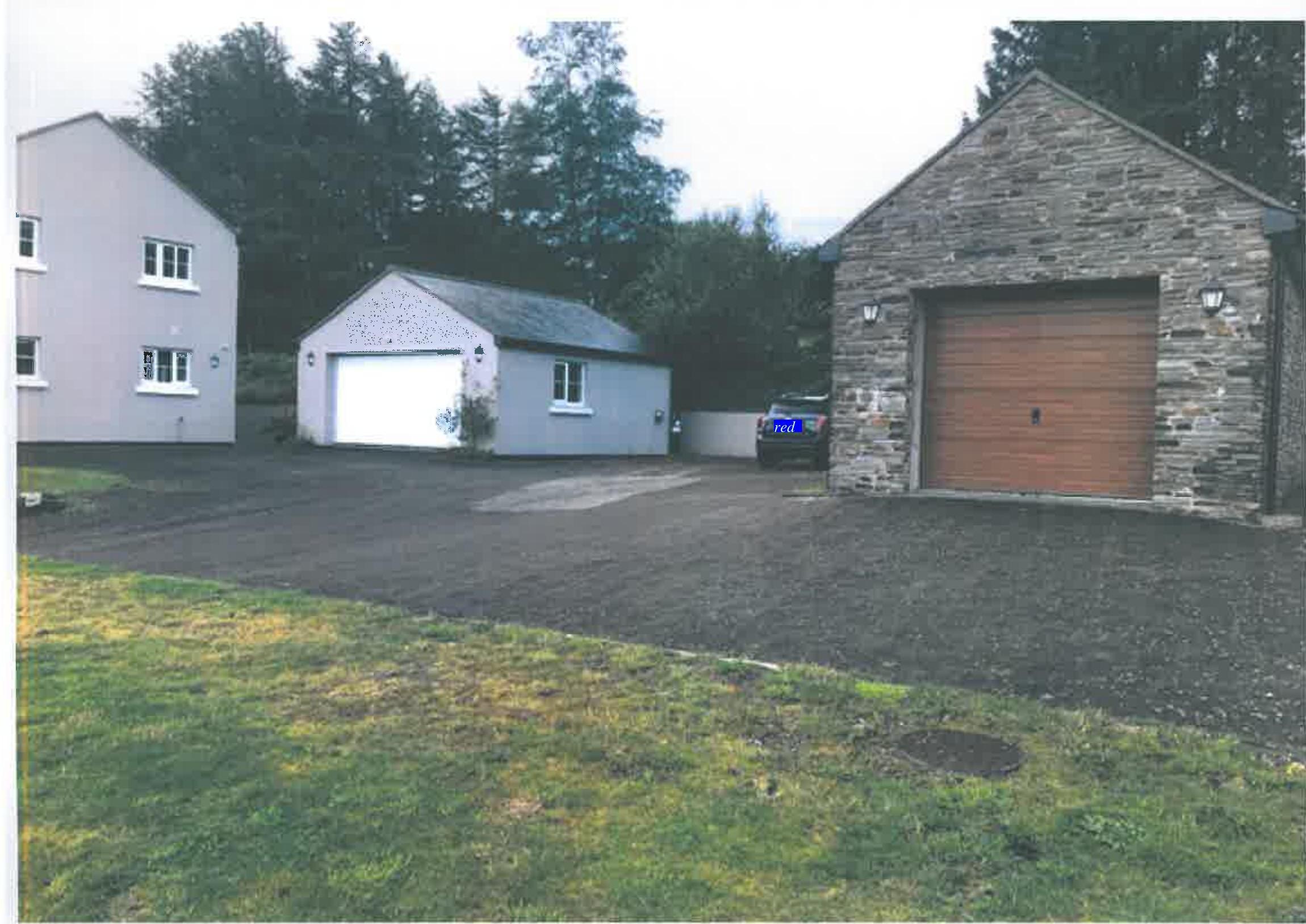 A photograph showing a residential property featuring a white two-story house, a detached garage, and a stone outbuilding with a large wooden door, all situated around a gravel driveway.