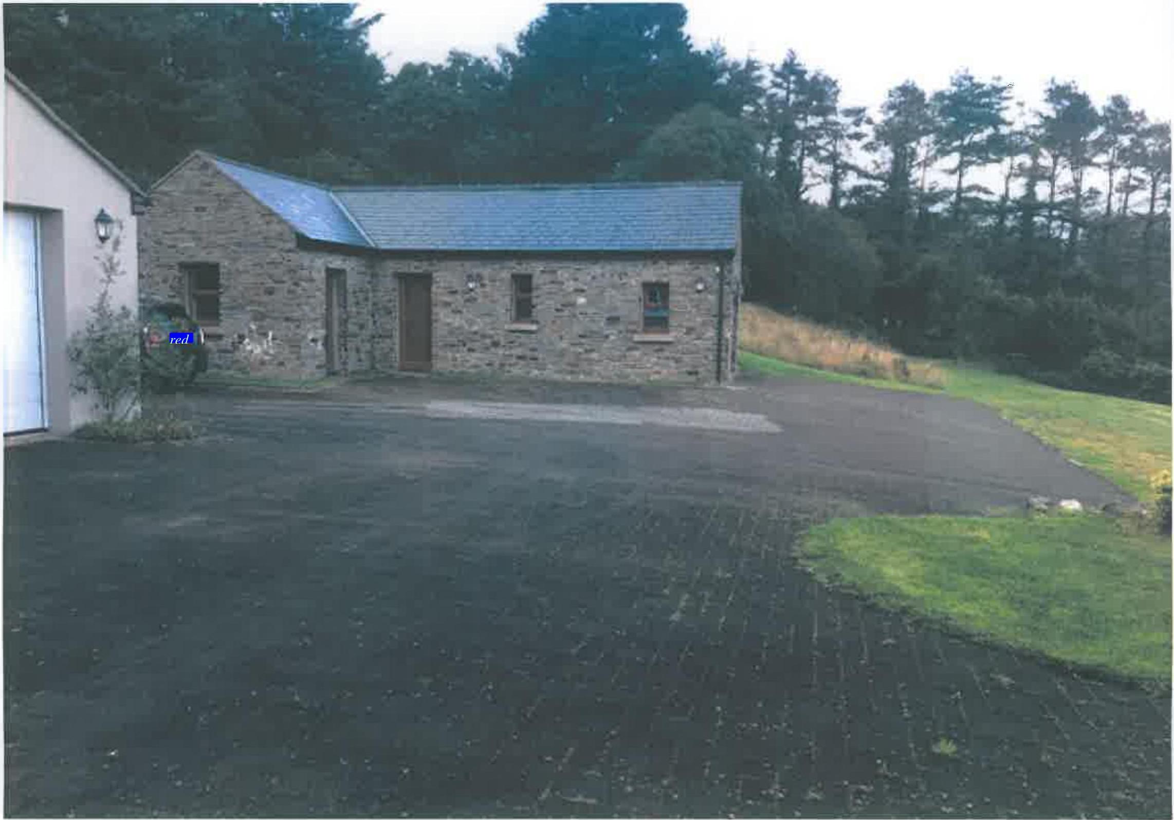 A photograph showing a stone single-story building with a slate roof, likely a converted barn, situated next to a white garage building. A large paved driveway is in the foreground with trees and a grassy slope behind.