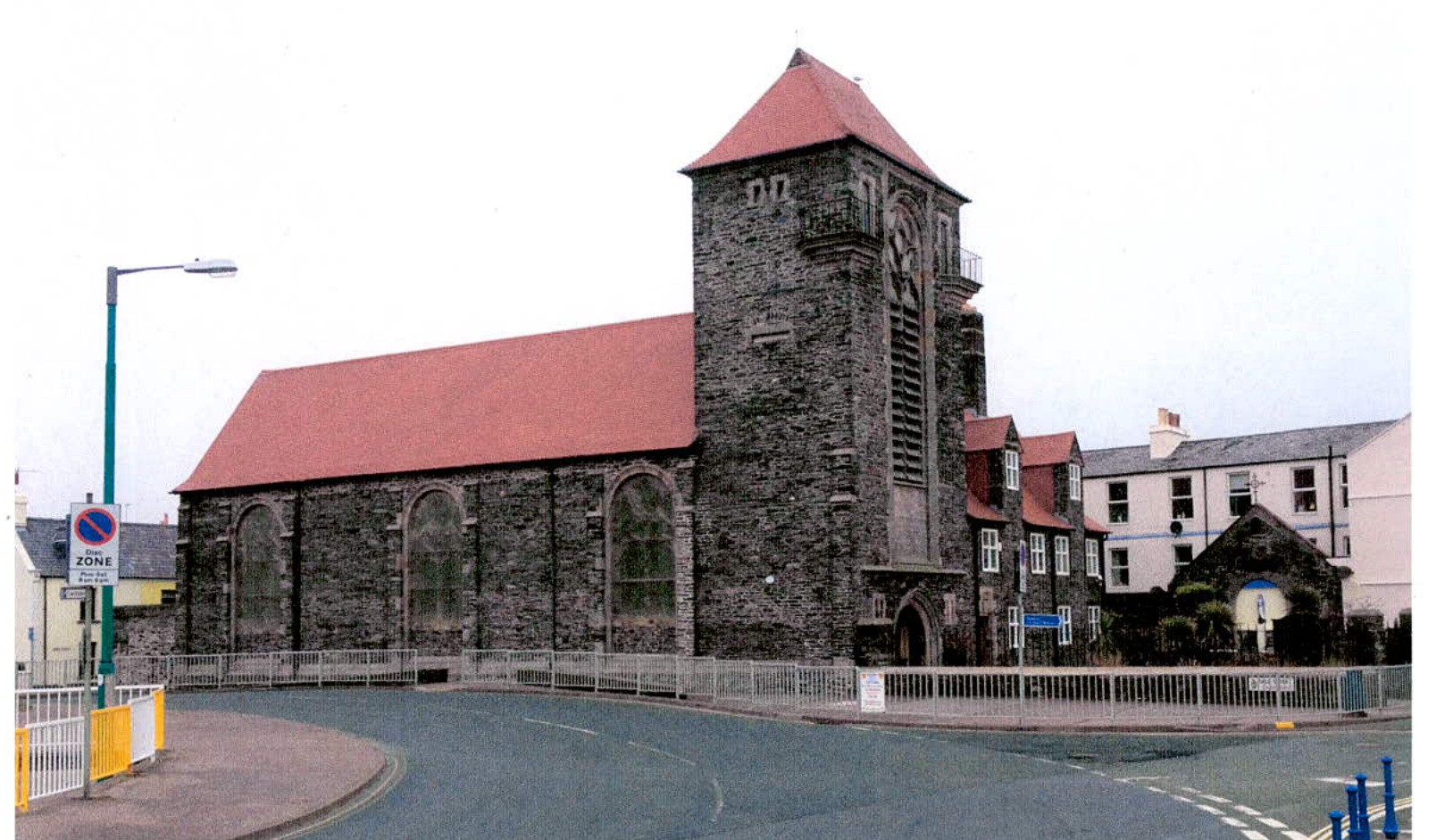 A photograph showing the exterior of a large stone building with a red roof and a prominent tower, situated next to a road.