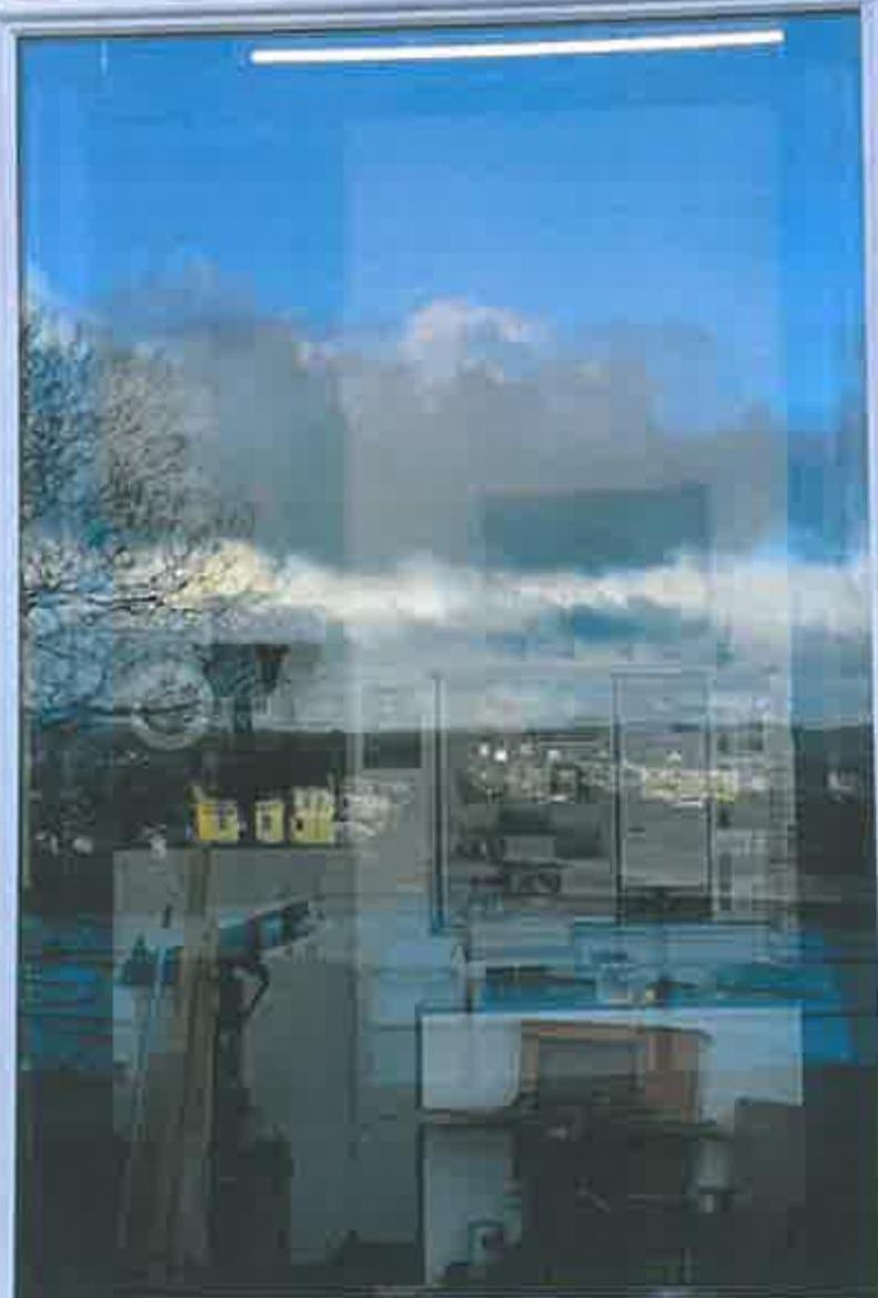 A close-up photograph of a window pane showing strong reflections of the blue sky, clouds, and nearby trees or buildings.
