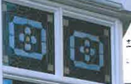 A close-up photograph showing two white-framed windows featuring decorative leaded glass panels with blue and yellow geometric designs.