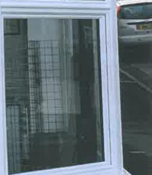 A close-up photograph of a white uPVC window frame with reflections visible in the glass. A car is partially visible in the background on the right side.